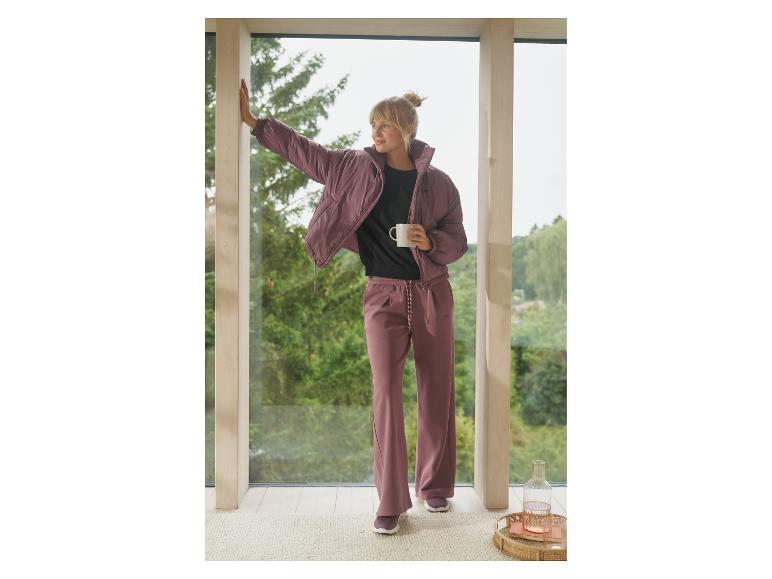 Vrouw in paarse pufferjas en bijpassende broek staat bij raam met een mok.