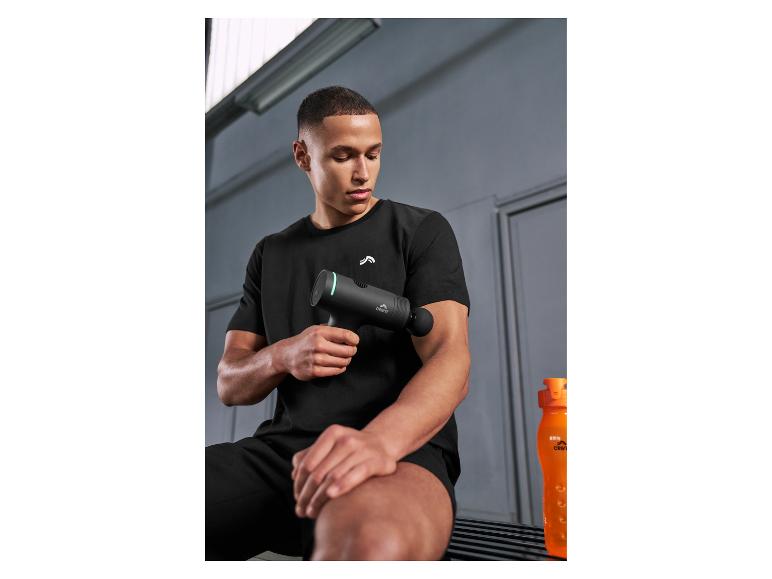 Man in zwarte sportkleding gebruikt een massagepistool op zijn arm, met een oranje waterfles in de buurt.