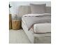 Beige bed met grijs beddengoed en kussen, met houten nachtkastjes en lamp ernaast.