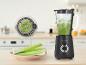 Een zwarte blender met een groene smoothie en selderij op een houten plank.