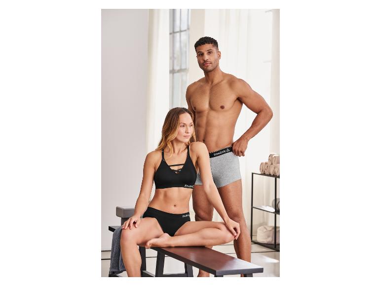 Man in grijze boxershort en vrouw in zwarte sportbeha en slip in een lichte kamer.