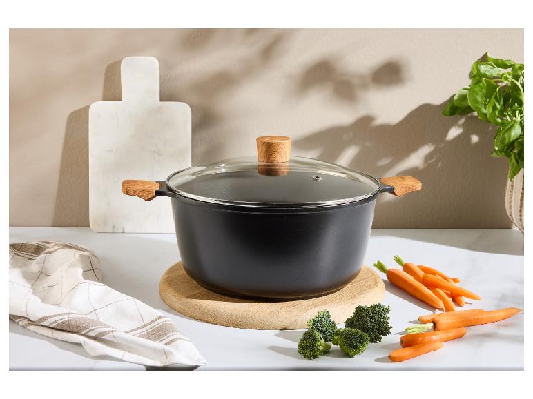 Zwarte kookpot met houten handvatten en glazen deksel, met groenten op het aanrecht.