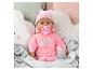 Babypop in roze outfit met speen en fles, omringd door zachte kussens.