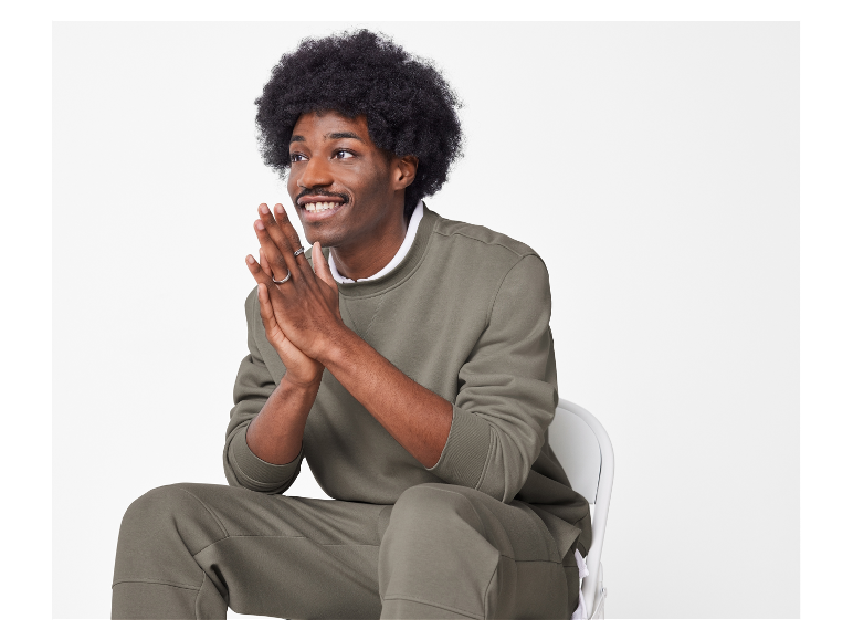 Man met afro zit op een stoel in een olijfgroene sweater en joggingbroek.