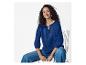 Vrouw in blauwe geborduurde blouse en jeans, zittend op een stoel, lachend.