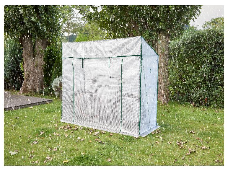 Een transparante kas in een tuin met bomen en gras, met regen die valt.