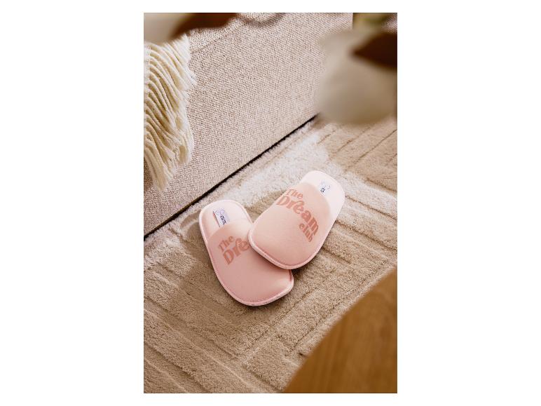 Roze 'The Dream Club' pantoffels op een beige vloerkleed naast een bank.