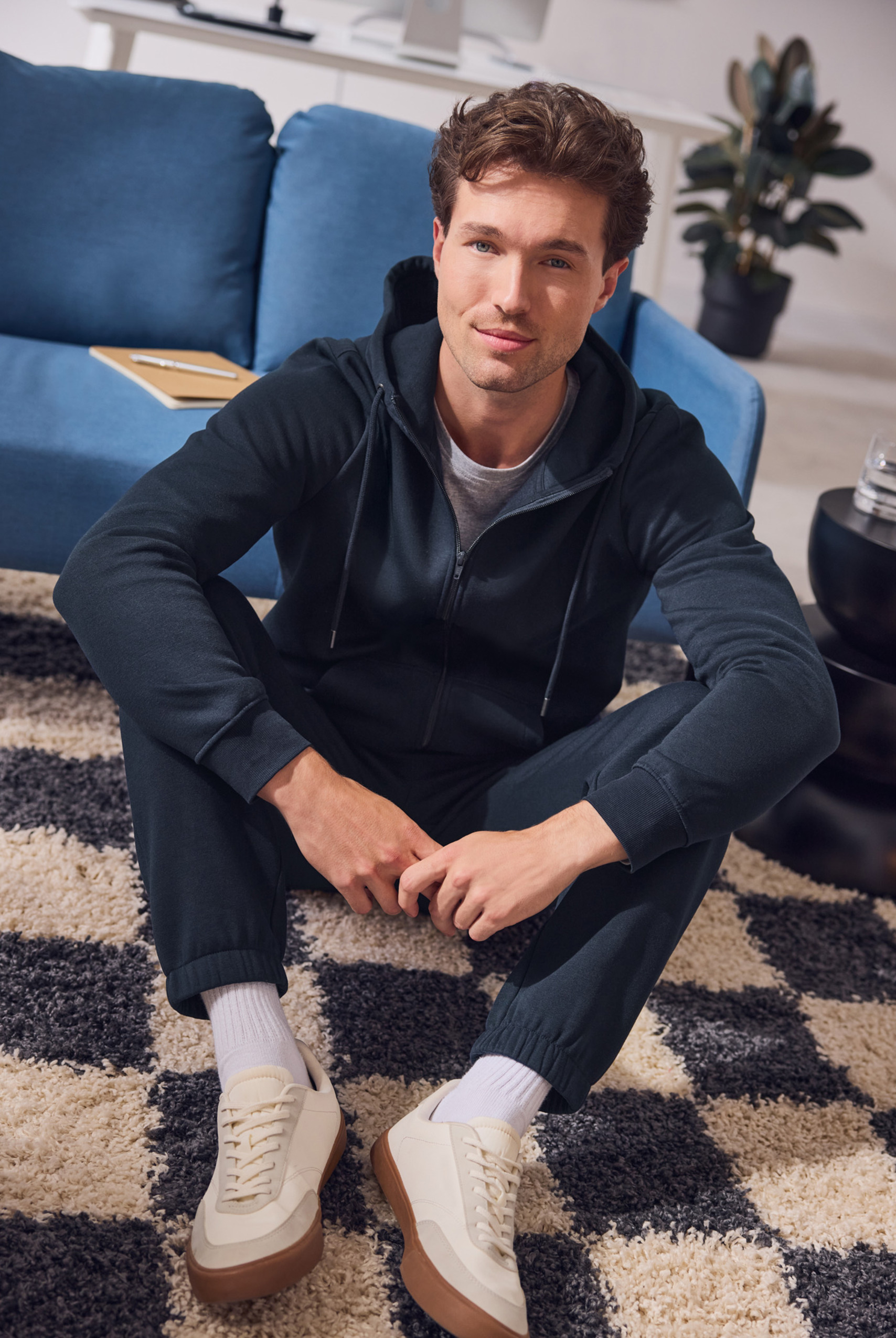 Man in een donkerblauwe hoodie en joggingbroek zit op een geruit vloerkleed.