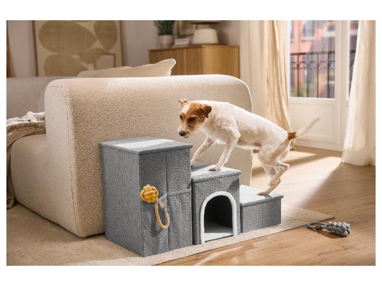 Hond beklimt grijze huisdierentrap met ingebouwd bed, naast een beige bank in een woonkamer.