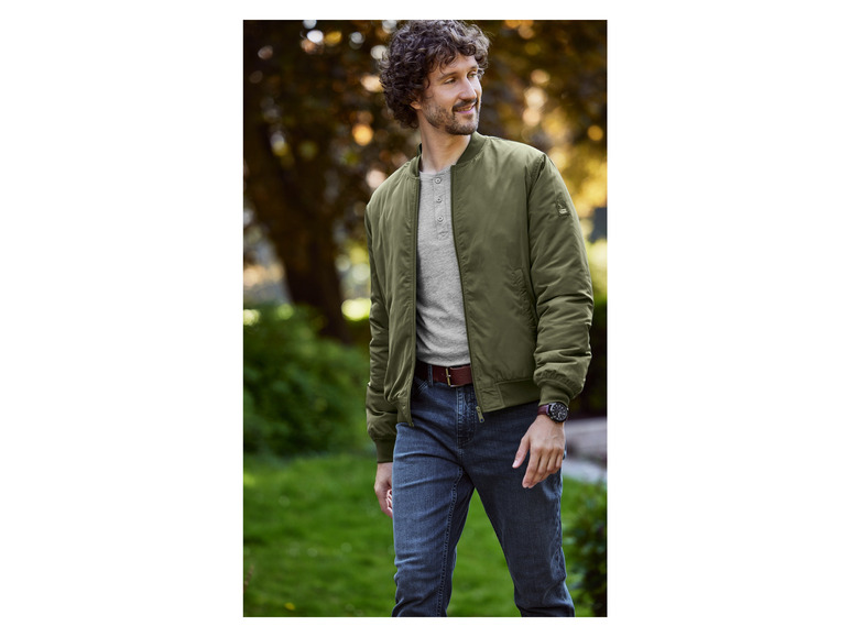 Man draagt een groene bomberjack, jeans en een horloge.