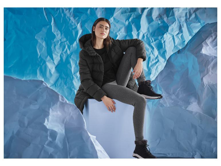 Vrouw in zwarte jas en grijze legging, zittend op een blauwe kubus, tegen een blauwe achtergrond.