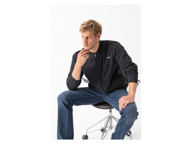 Man in zwart bomberjack en blauwe jeans, zittend op een stoel.