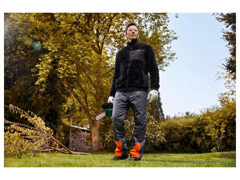 Een man in een tuin met een zwart PARKSIDE fleecevest en een werkbroek.