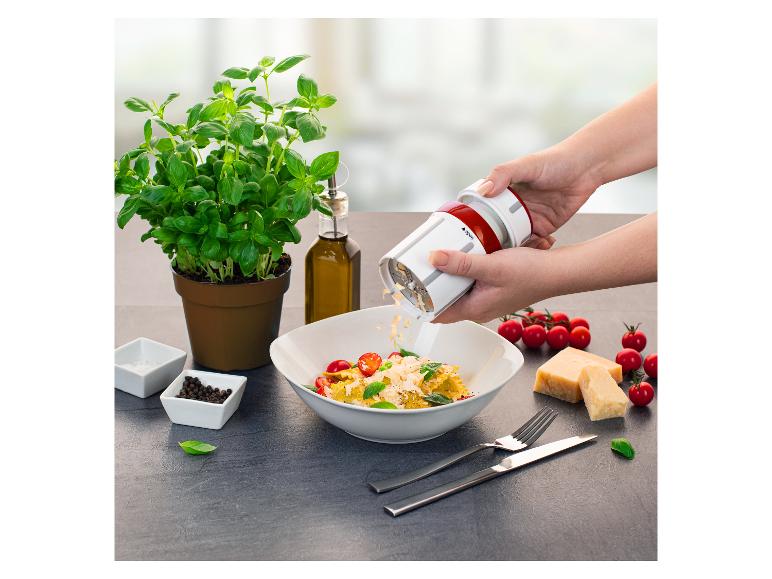 Een persoon raspt kaas over een bord pasta met cherrytomaten en basilicum, met olijfolie en kruiden.