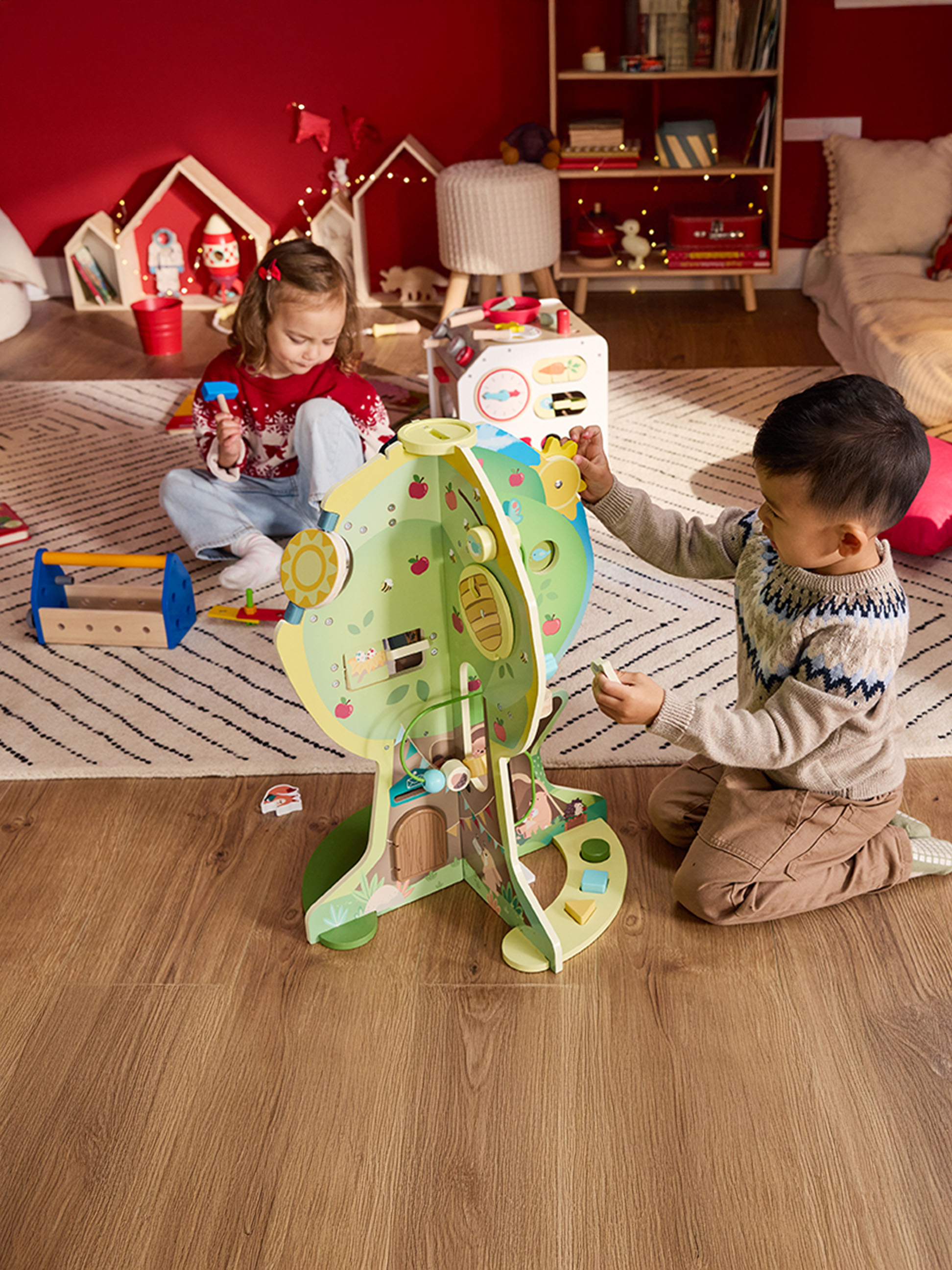 Twee kinderen spelen met een houten poppenhuis en een gereedschapskist in een feestelijk versierde kamer.