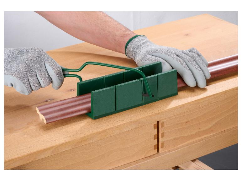 Handen in handschoenen gebruiken een verstekbak en zaag om een stuk lijstwerk op een houten werkbank te zagen.