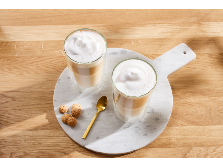 Twee glazen gelaagde latte macchiato met schuim, een gouden lepel en koekjes op een marmeren plank.