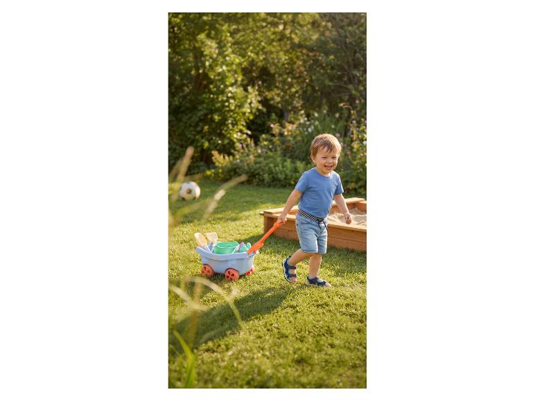 Lachende jongen in blauwe korte broek en T-shirt, die een speelgoedkarretje trekt in de tuin.