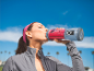 Vrouw drinkt rode smoothie uit draagbare blender, buiten onder een blauwe lucht.