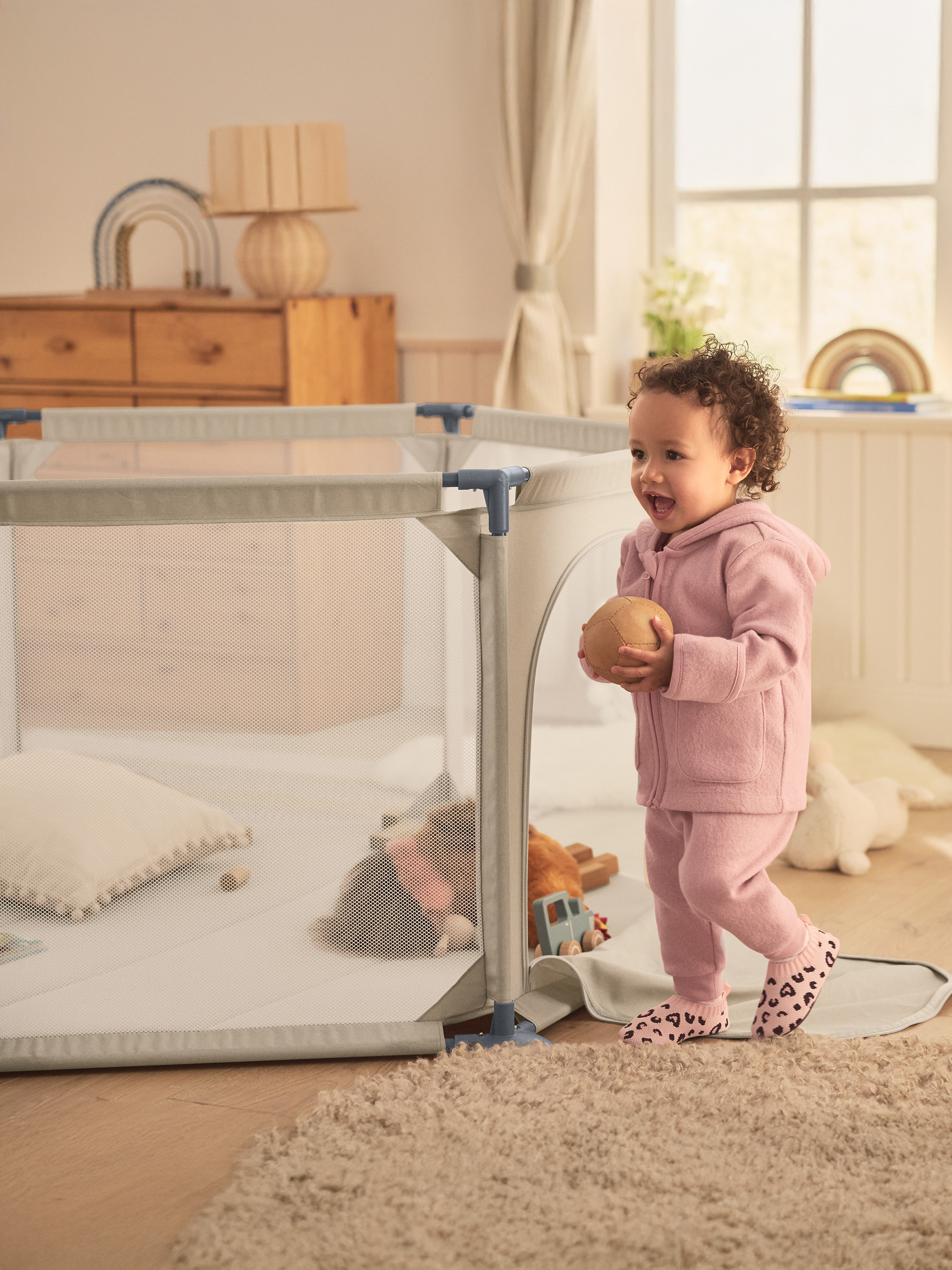 Peuter in roze outfit met bal, staand naast een box met speelgoed erin.