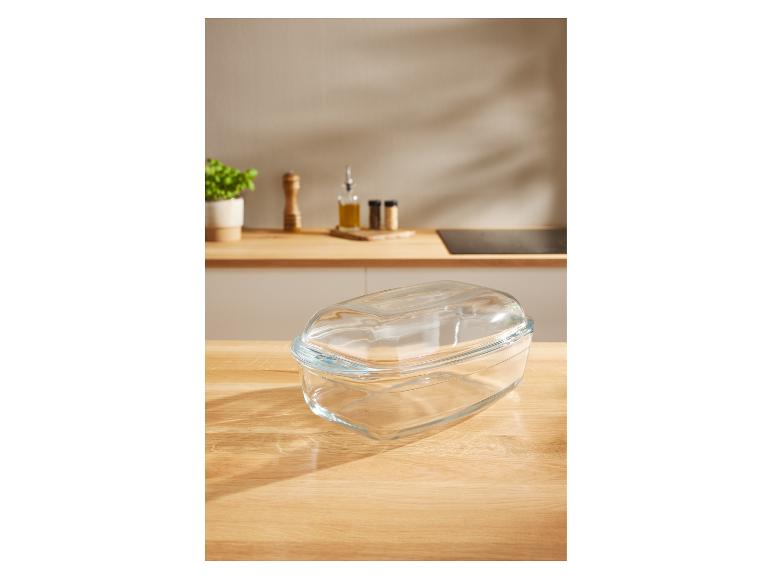 Glazen ovenschaal met deksel op een aanrecht met kruiden en een plant op de achtergrond.