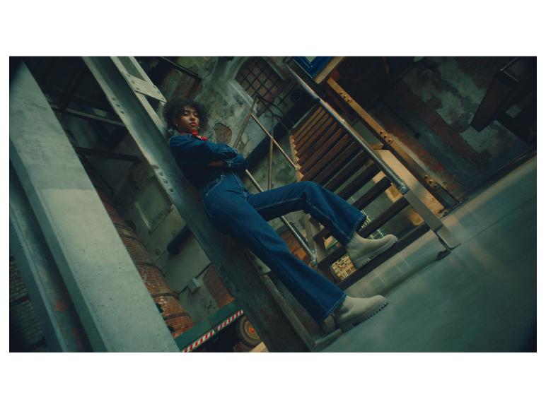 Een vrouw in een denim jumpsuit en lichte laarzen poseert op industriële trappen.