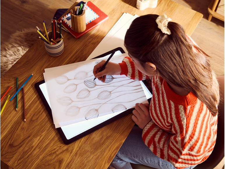 Vrouw tekent op een lichttafel met kleurpotloden en notitieboekjes op een houten tafel.