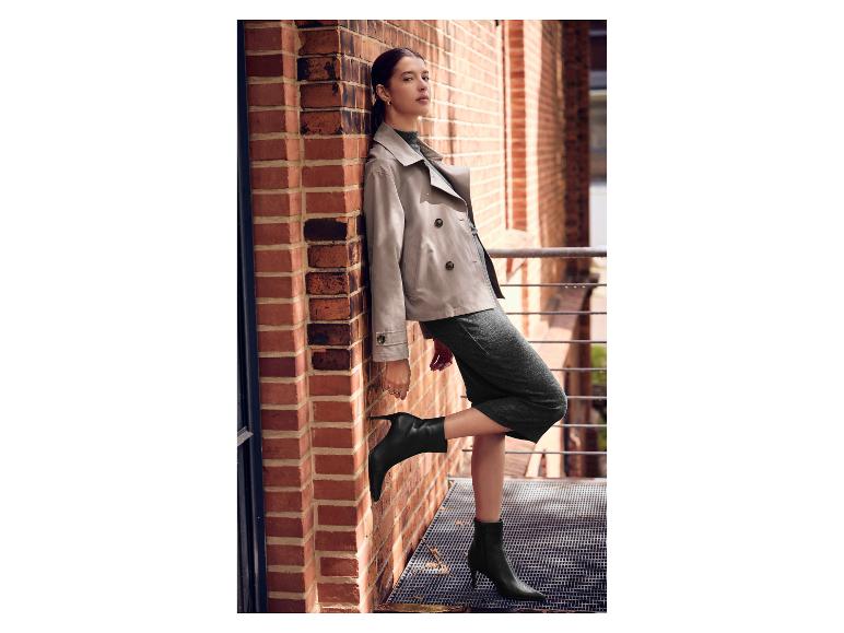 Vrouw in beige trenchcoat en grijze jurk, met zwarte hakken.