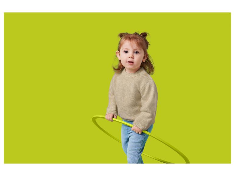 Een peuter met roodachtig haar, in een beige trui en spijkerbroek, houdt een groene hoepel vast.