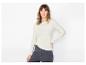 Vrouw in een lichtgekleurde blouse met lange mouwen en donkere jeans, poserend tegen een witte achtergrond.