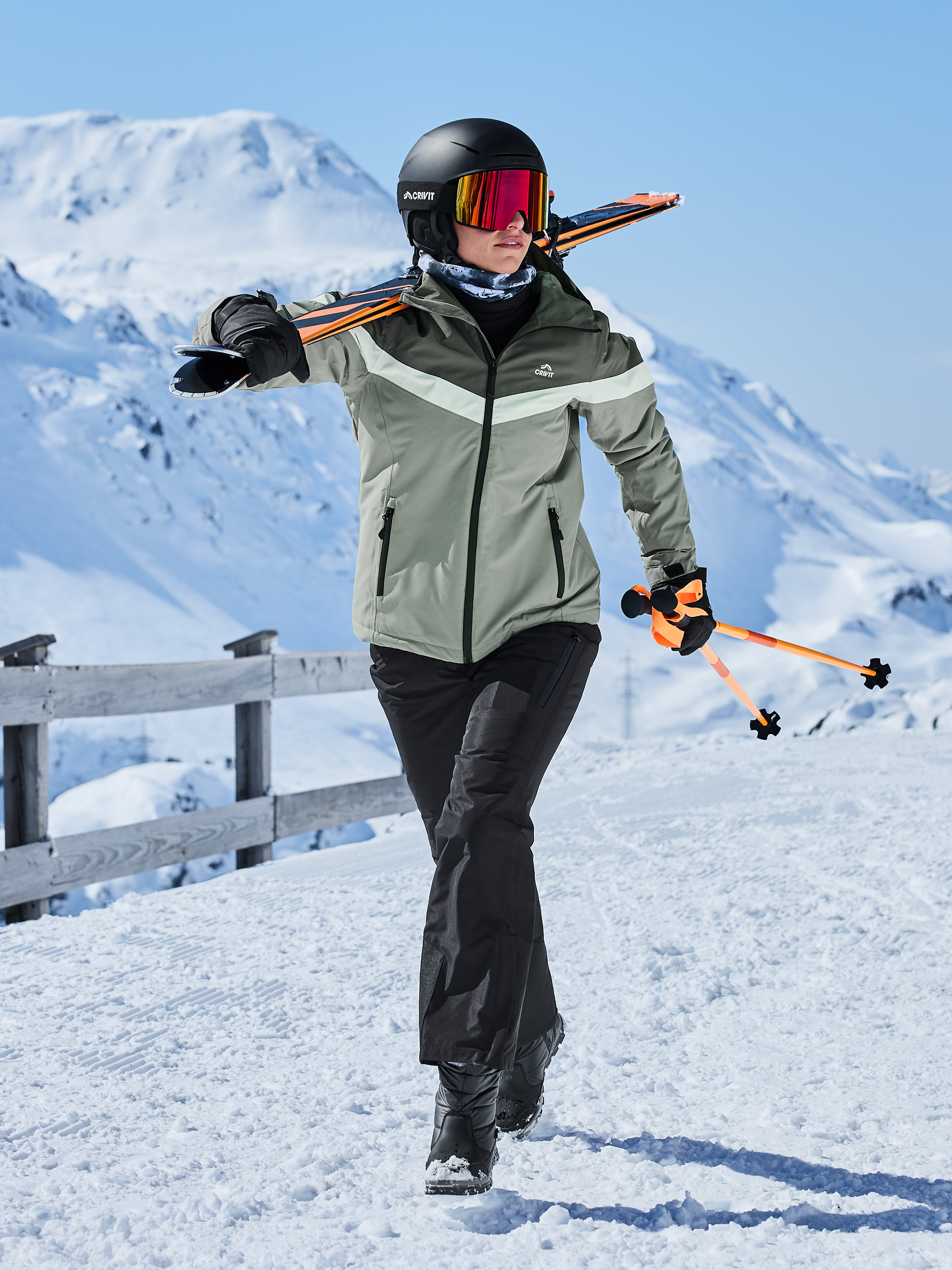 Vrouw in ski-jas, broek, helm, skibril en ski's over schouder in de sneeuw.