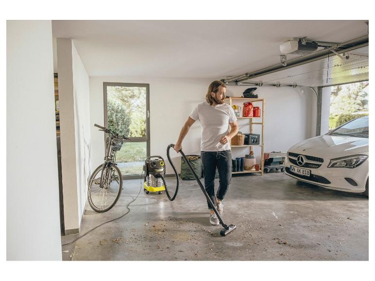 Man stofzuigt garage met een nat- en droogzuiger, met fiets en auto op de achtergrond.