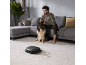 Man speelt videogames met hond en robotstofzuiger op een licht tapijt.