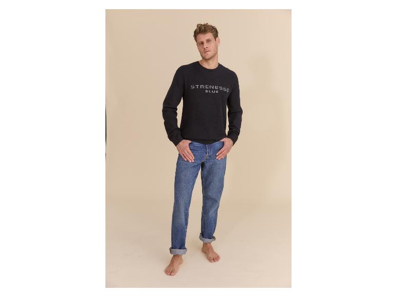Man in een zwarte trui met logo en blauwe jeans, op blote voeten.