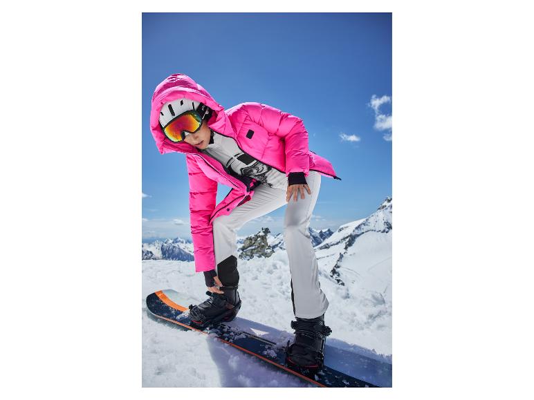 Een vrouw in een roze jas en witte snowboardbroek, die bindingen op een snowboard aanpast.