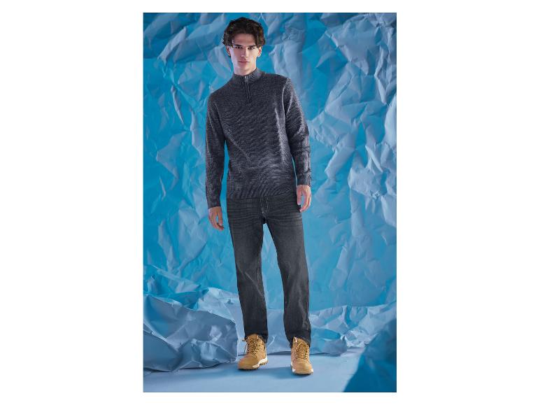 Een man in een grijze trui en donkere jeans voor een blauwe achtergrond.