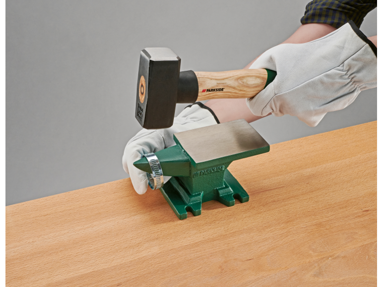 Een hand in een werkhandschoen houdt een hamer boven een klein groen aambeeld op een houten tafel.