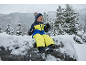 Kind in geel-blauw skipak, muts en handschoenen, zittend op een besneeuwde rots.