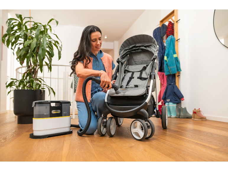 Een vrouw reinigt een kinderwagen op een houten vloer met een tapijtreiniger.