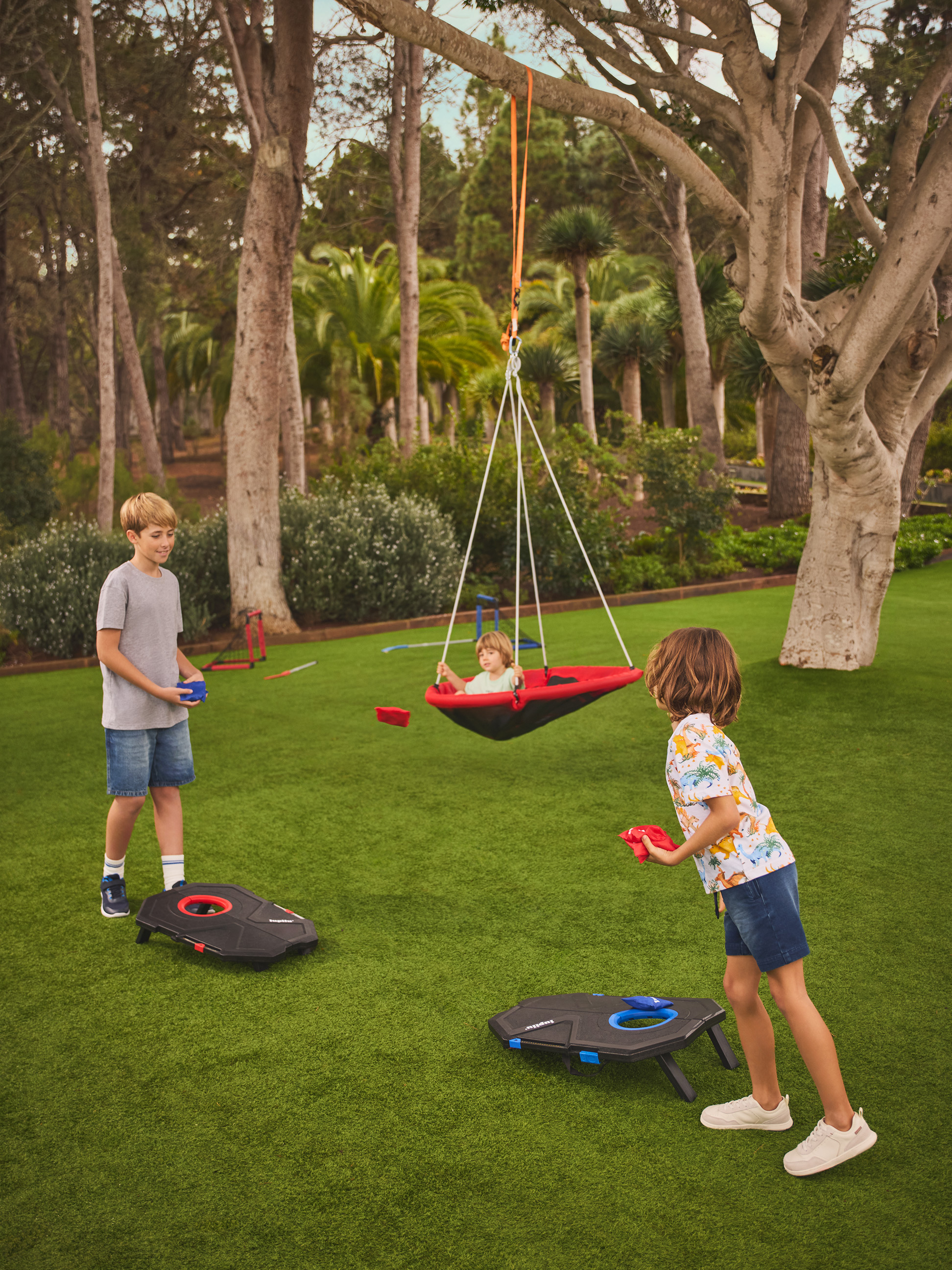 Twee kinderen spelen cornhole op het gazon, terwijl een derde in een nestschommel zit.