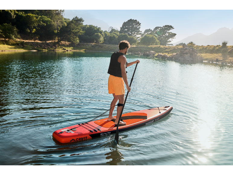 Man peddelt op een paddleboard op een meer, omringd door bomen en bergen.