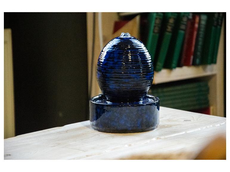 Blauwe keramische binnenfontein op een houten tafel, met boeken op de achtergrond.
