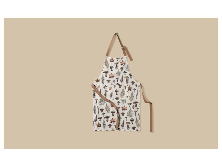 Schort met paddenstoel- en varenpatroon, hangend aan een haak tegen een beige muur.