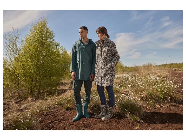 Een stel in regenjassen en regenlaarzen staat voor groene bomen en een blauwe lucht.