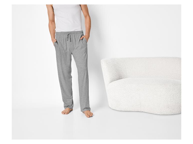 Man in grijze joggingbroek en wit hemd, staand naast een witte fauteuil.