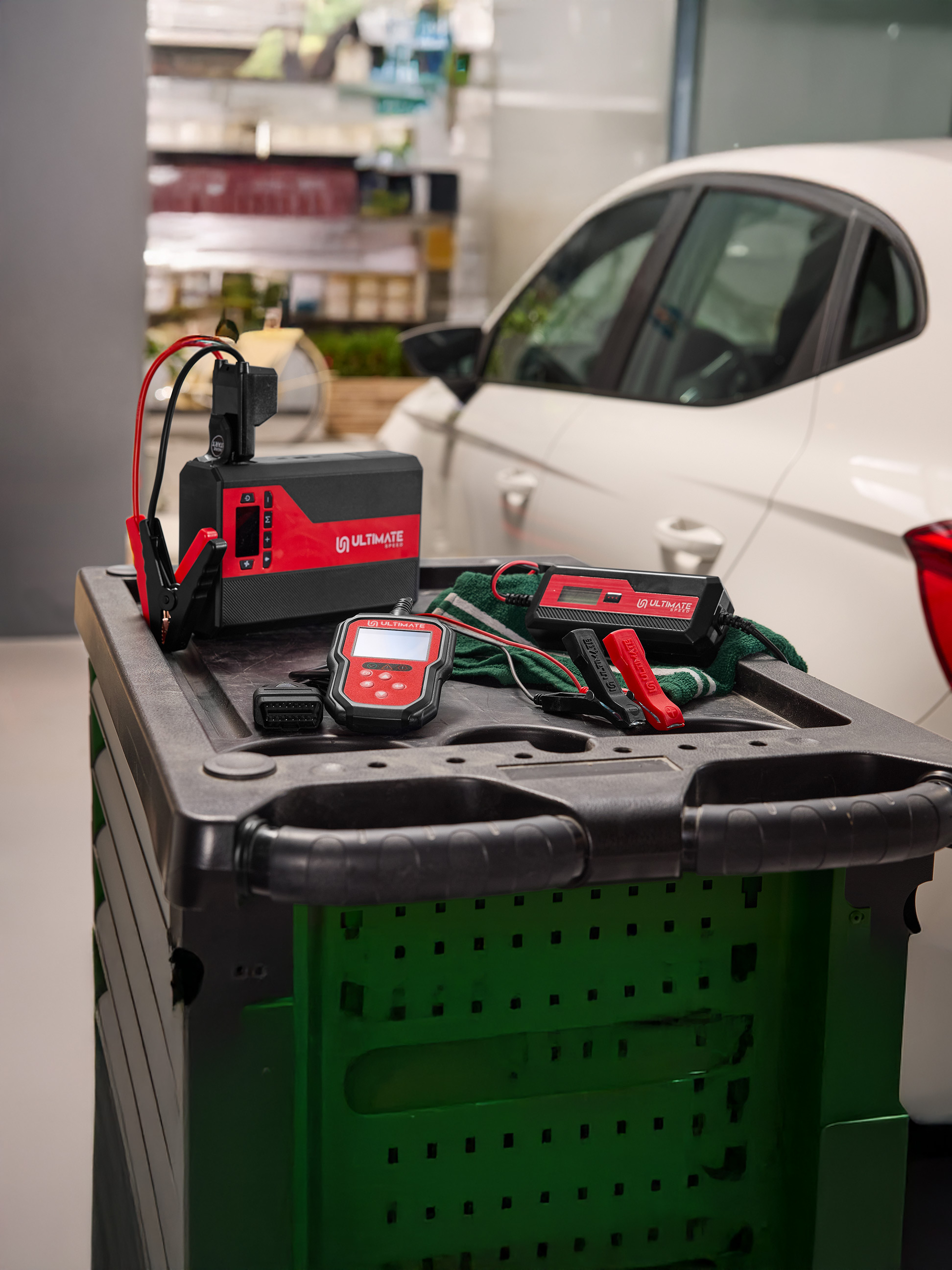 Autobatterijlader en diagnostische tools op een kar naast een witte auto.