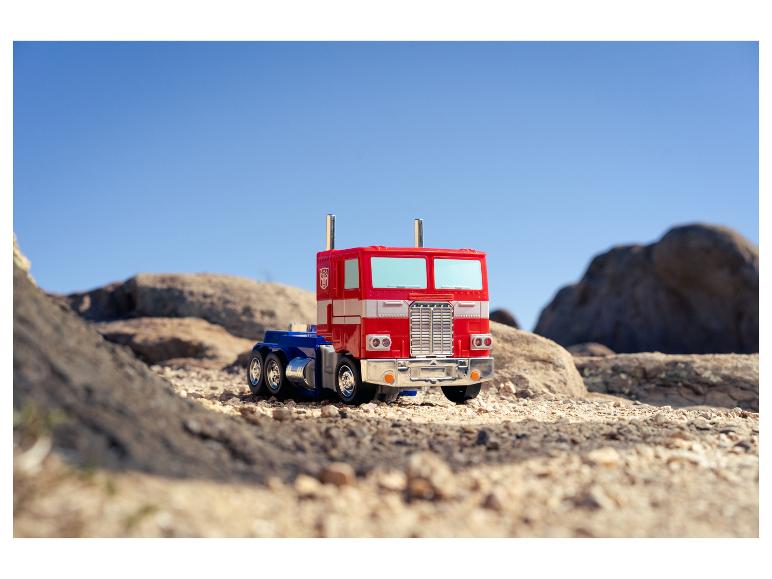 Een rode en blauwe Optimus Prime speelgoedtruck op rotsachtig, zanderig terrein onder een heldere hemel.