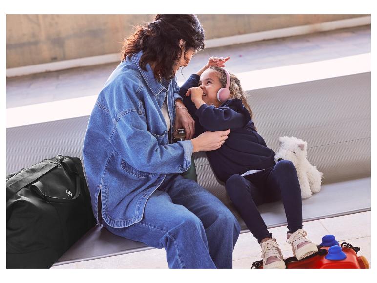 Een moeder en dochter zitten op een bank met een zwarte reistas en een kinderkoffer.