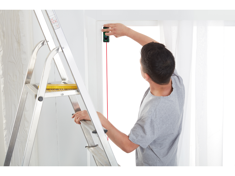 Man op een ladder gebruikt een laserafstandsmeter om een kozijn te meten.