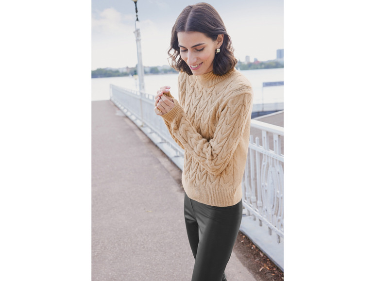 Een vrouw draagt een beige kabeltrui en zwarte broek